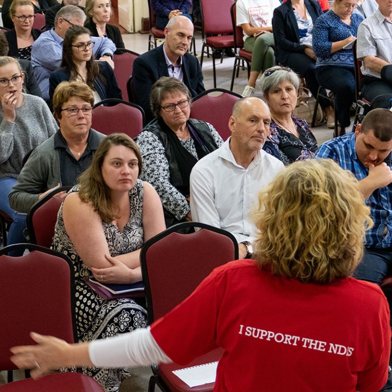 Make it Work audience members looking at Kirsten our MC in the foreground, with an arm outstretched wearing a red EAC teeshirt that reads "I support the NDIS"