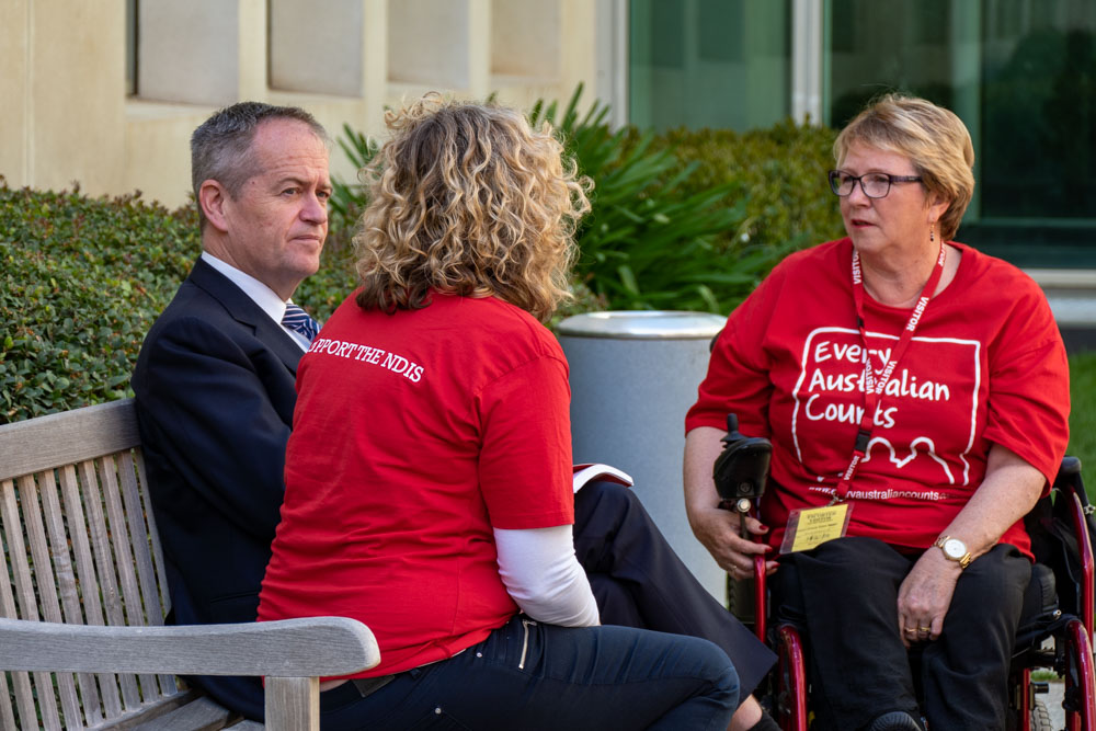 Bill Shorten MP looking at EAC Campaign Director Kirsten Deane, who are both on a park bench. Kirsten's back is facing us. The back of her teeshirt reads "I support the NDIS". EAC Champion Lynne Foreman is sitting across from them, also listening. They are in a garden at Parliament House. 