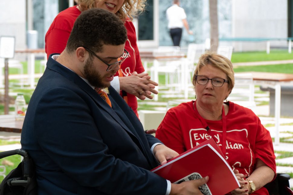 Senator Jordon Steele-John looking at the Messages to the Minister book. EAC Campaign Director Kirsten Deane is standing beside him looking over his shoulder at the book, but her head is cropped out. EAC Champion Lynne Forman is next to Kirsten, also looking at the book. They are in a courtyard at Parliament House. 