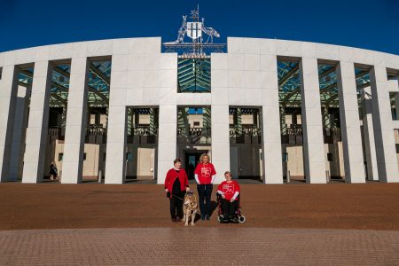 Leah and her guide dog Lisa, and Kirsten and Lynne standing outside Parliament House on a bright day.