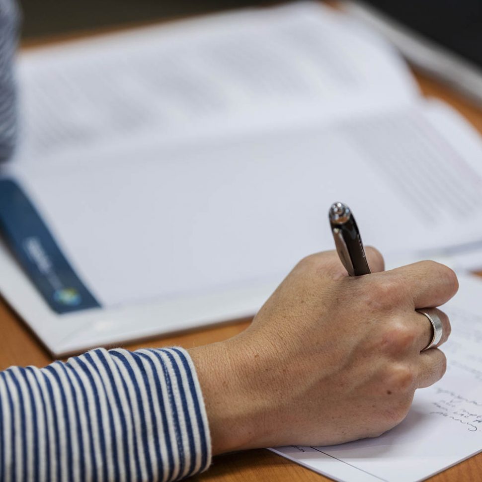 Photo of a woman's hand, holding a pen and writing. Out of focus paperwork is in the background.