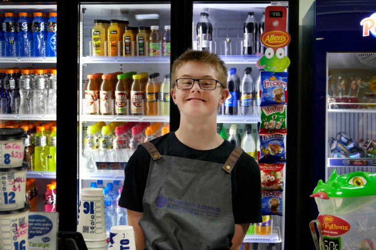 Julian standing behind the counter with a drinks fridge behind him. He is wearing a black teeshirt and an apron