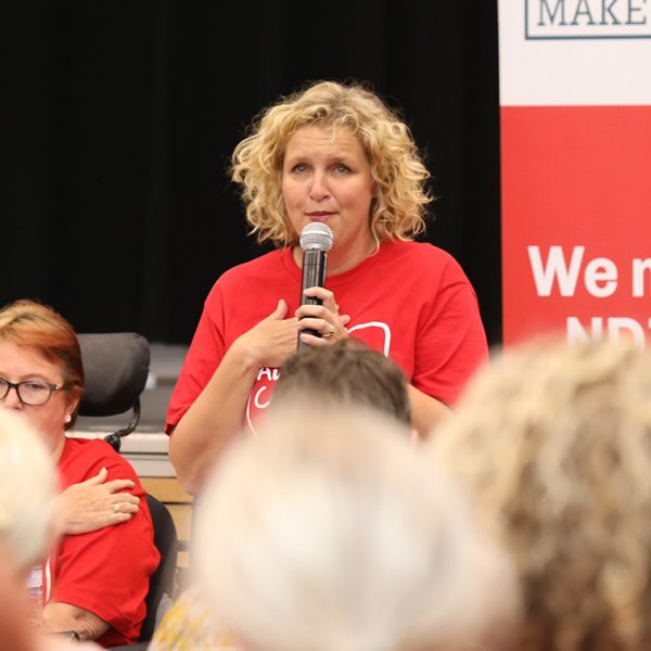 EAC Campaign Director Kirsten Deane holding a mic with her other hand on her chest in a town hall, with lots of people around