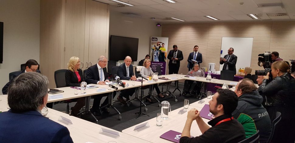 People with disability and families in a meeting room around a series of tables in a big rectangle. The media is there, and so are people from the government; Prime Minister Scott Morrison, Minister for the NDIS Stuart Robert, NDIA CEO Vicky Rundle, and more. 