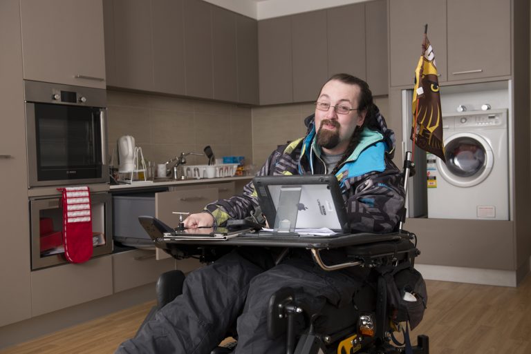A man using a motorised wheelchair, with an iPad, in his accessible apartment.