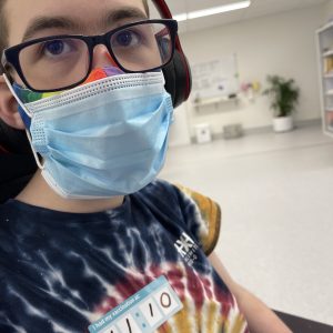 Photo of Bodhi - a teenage wheelchair user in a medical clinic. He has glasses, a rainbow mask, a surgical mask, and a sticker that reads "I got vaccinated at 11:10".