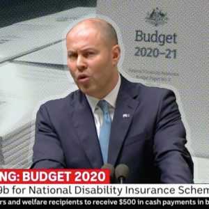 Still of Treasurer Josh Frydenburg giving his budget speech in Parliament, with printed copies of the 2020-21 Budget behind him. The ABC News ticker tape below says "Breaking: Budget 2020. Extra $3.9B for National Disability Insurance Scheme.