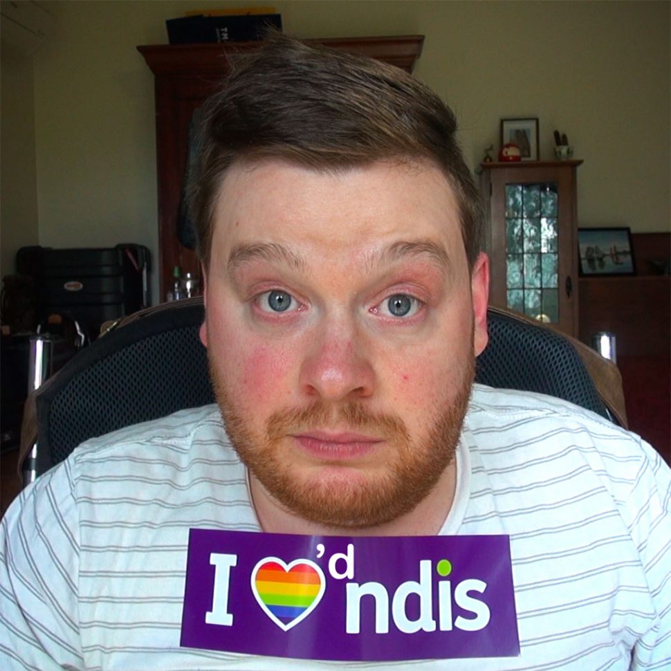 Carl Thompson, a man using a wheelchair in his living room. He has a sticker on his chest that he has modified so that it reads "I loved NDIS" instead of "I love NDIS"