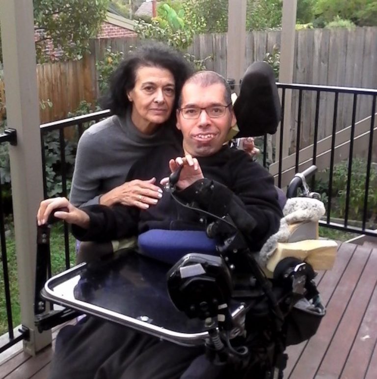 Photo of Dr George Taleporos and his mum Kathy on the back deck of a house. Dr George uses an electric wheelchair, and his mum is standing behind him with a hand across his arm and chest.