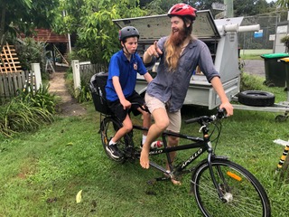 A teenage boy on the back of a tandem bike, behind a young man with a big beard. They're in a very tropical looking front yard, about to start riding.