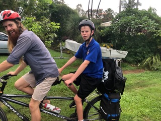 Two young men sharing a tandem bicycle with bags on the back, in a tropical front yard