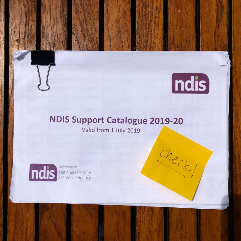 Photo from above of the 19-20 NDIS Support Catalogue print out. It's held together with a big bulldog clip, and has a yellow sticky note on it reading 'check!'. It's on a wooden outdoor tabletop.