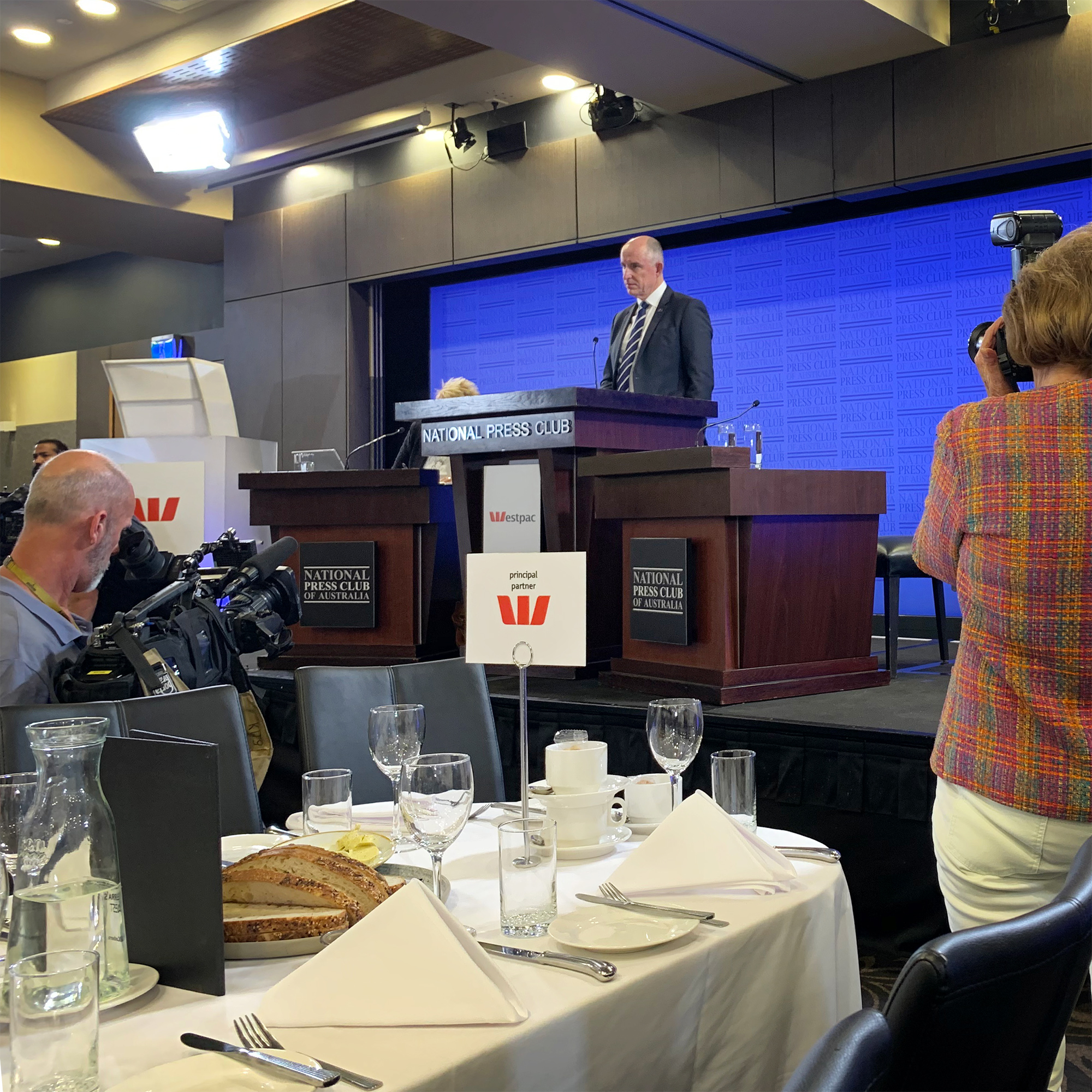 Photo of Minister for the NDIS Stuart Robert MP, standing behind the National Press Club podium on a stage in the middle. In the foreground is a table with plates, glasses, cutlery and bread. There is a cameraman recording on the left, and a photographer on the right.