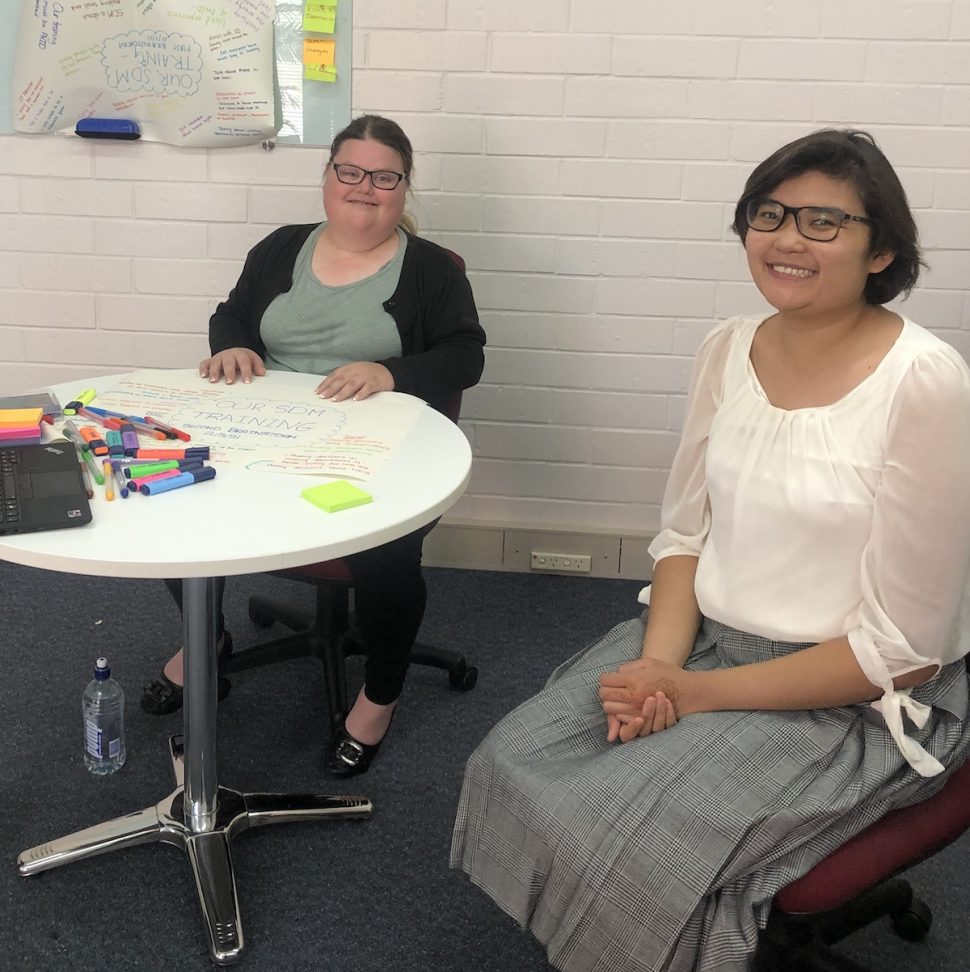 Two young women with intellectual disabilities smiling, sitting on office chairs by a table with butchers paper, sharpies and sticky notes.