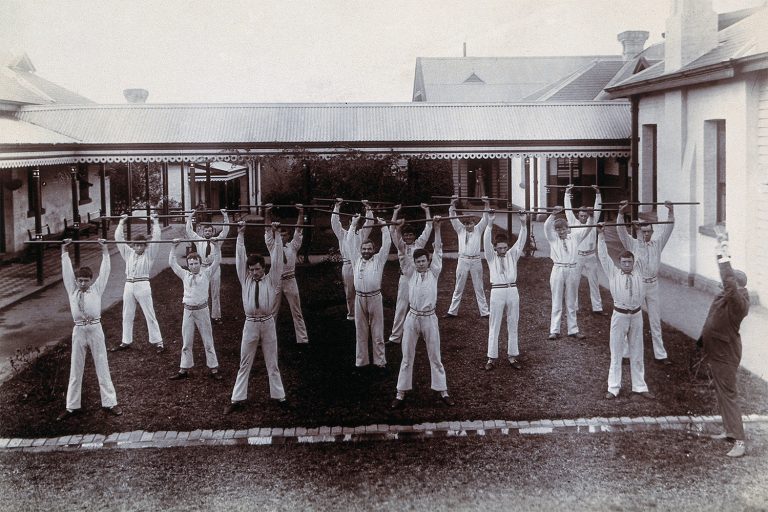 Black and white photo from the late 1800’s showing a group of mean wearing white uniforms, exercising in a courtyard at what was formerly called Metropolitan Lunatic Asylum, in Kew, Victoria.