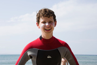 A young man with light curly hair, a big smile, and white zinc on his face standing on a beach on a sunny day.