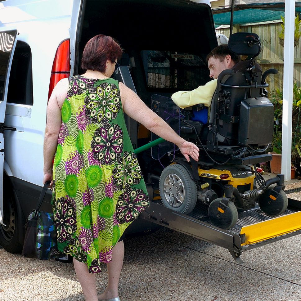 A woman in a green dress points backwards as a man using a motorised wheelchair reverses onto the ramp lift at the back of a van.