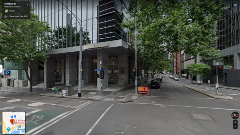 Grey building on a corner, with grey pillars. It is at an intersection of traffic lights. There are ramps to access the street/footpath. 