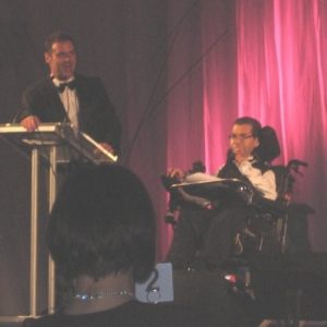 Dr George Taleporos on stage at an event. He is using an electric wheelchair. A celebrity is wearing a tuxedo at the podium in front. There are red curtains in the background.