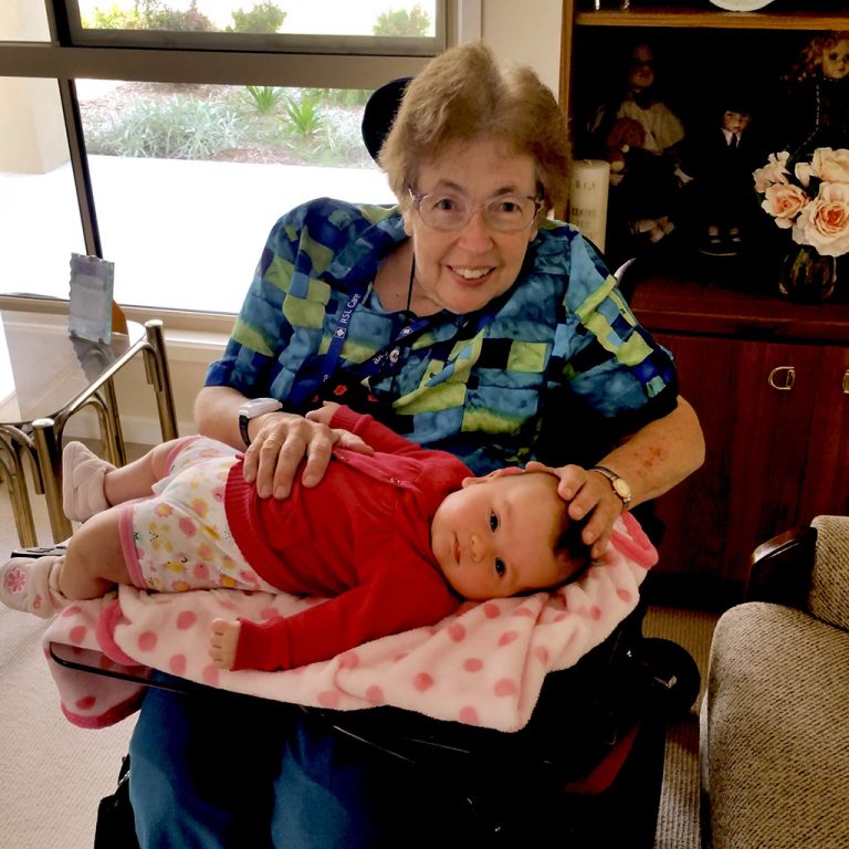 Heather, sitting in her wheelchair in a living room, smiling while holding her great niece, a gorgeous baby.
