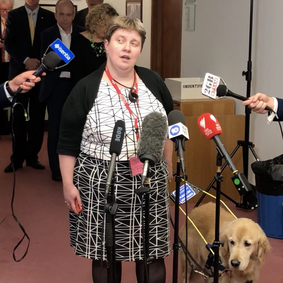 Photo of Leah van Poppel, Chair of the National Disability and Carer alliance speaking at Parliament House with lots of microphones in front of her, and her Guide Dog Lisa by her side.