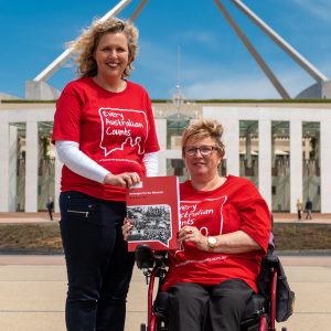 Photo of EAC Campaign Director Kirsten Deane, and EAC Champion Lynne Foreman out the front of Parliament House in Canberra. Kirsten is standing and Lynne is using a wheelchair. The are holding up a thick A4 book - the EAC messages for the Minister.