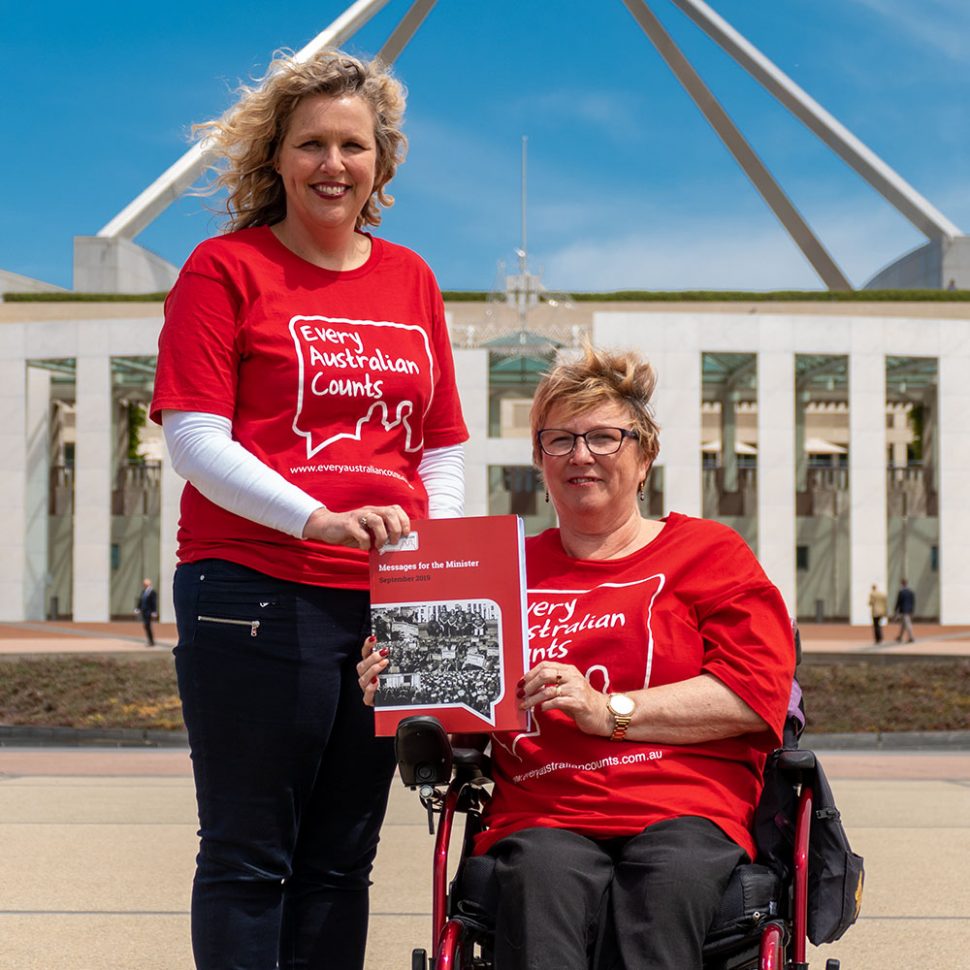 Photo of EAC Campaign Director Kirsten Deane, and EAC Champion Lynne Foreman out the front of Parliament House in Canberra. Kirsten is standing and Lynne is using a wheelchair. The are holding up a thick A4 book - the EAC messages for the Minister.
