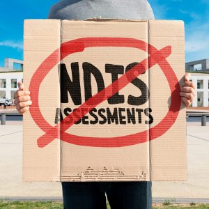 A protestor outside Parliament House holding a sign with NDIS assessments in a circle and crossed out.