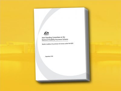 Parliament house in yellow, behind a copy of the Joint Standing Committee on the National Disability Insurance Scheme Market readiness for provision of services under the NDIS September 2018 report