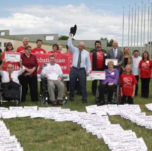 Every Australian Counts supporters on the lawn outside Parliament House in Canberra. They're holding signs up, wearing red EAC teeshirts, and on the lawn papers with the names of Australian cities and towns spell out NDIS. Tim Fischer stands in the centre of the group, raising his Akubra into the air.