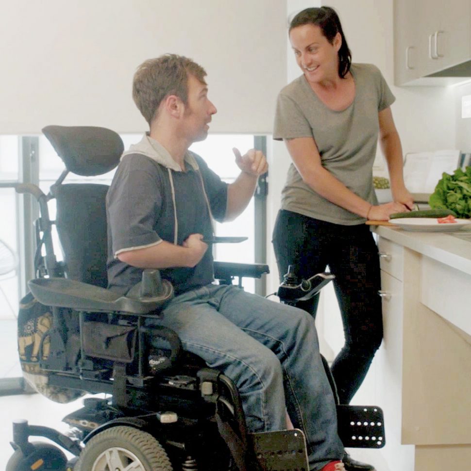 Two young adults in an accessible kitchen talking. The man is using a motorised wheelchair and the woman is standing.